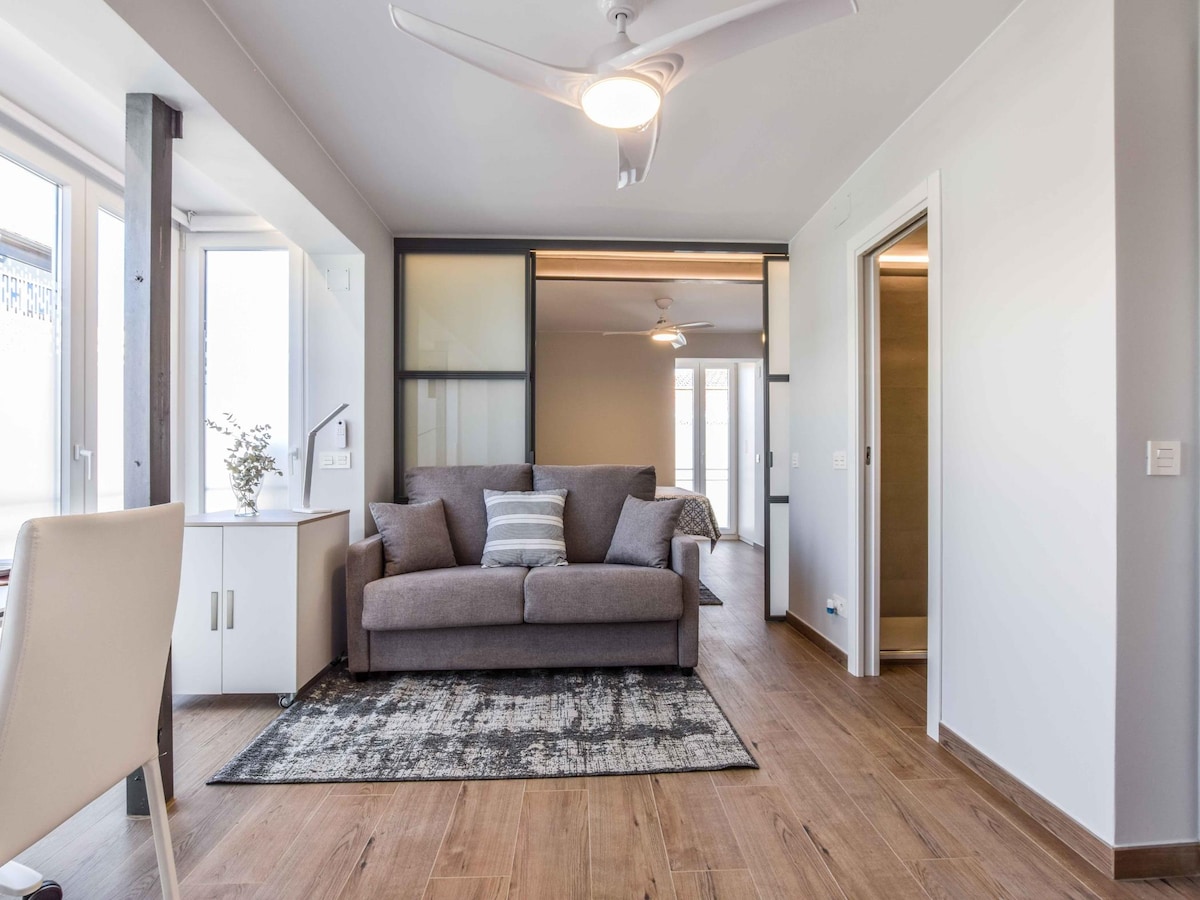 A light-filled living area features a comfortable gray sofa bed against a neutral backdrop. Natural light floods through two windows, while a simple rug defines the space. A ceiling fan is mounted overhead, and a solid divider leads to the bedroom area.