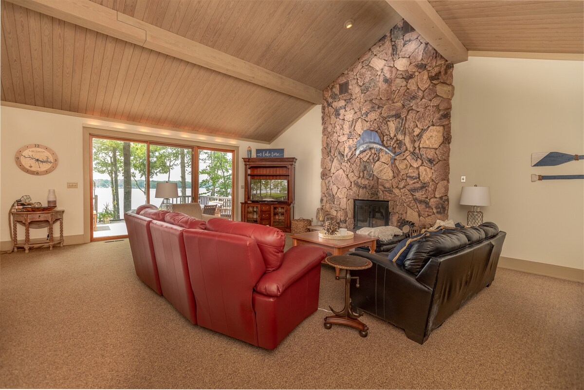 The inviting living area features a stone fireplace as its focal point, with large windows inviting natural light and lake views. Plentiful seating options are provided by two leather sofas in contrasting colors, complemented by a wooden side table for convenience.