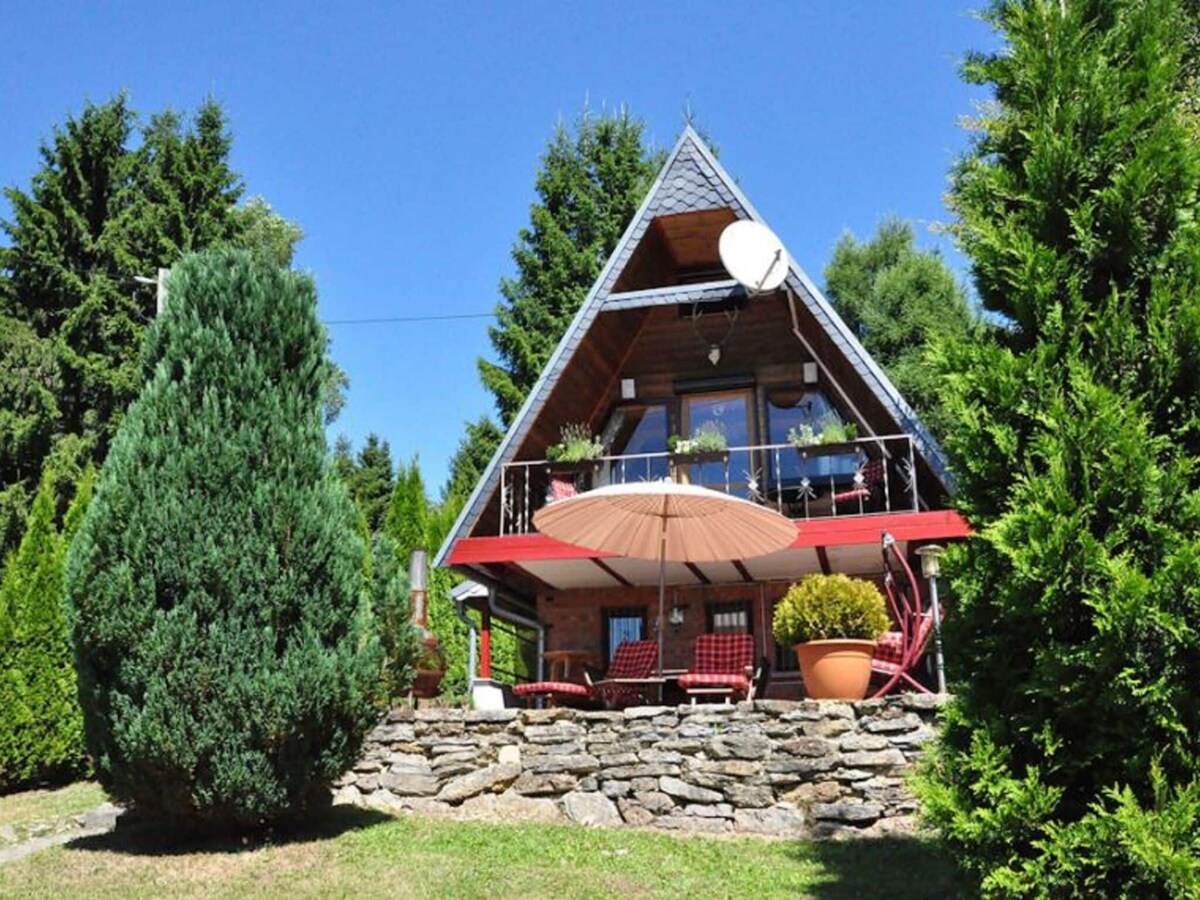The charming exterior of the holiday home is framed by lush greenery and tall trees. A red umbrella shades a small terrace, which features comfortable seating. The sloped roof adds a distinctive character to the house, creating a cozy and inviting appearance.