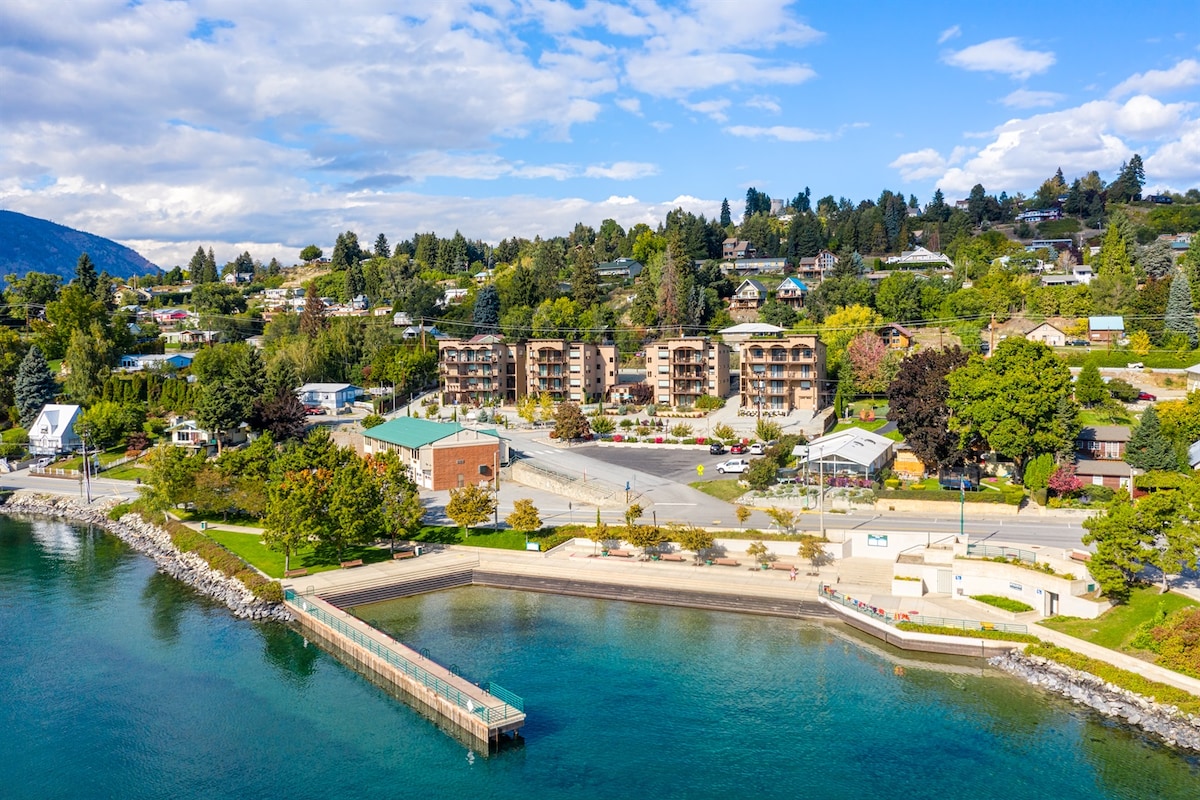 The image captures a scenic view of the Marina’s Edge condos, located near the shoreline. The multi-story building is surrounded by lush greenery and a paved area, with clear blue waters in the foreground and a dock extending into the lake.