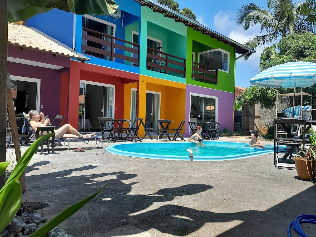 A vibrant shared space features a circular pool surrounded by lounge chairs. Colorful facades of the building add character to the setting. Palm trees provide shade, and an outdoor eating area is visible, enhancing the inviting atmosphere of the communal area.