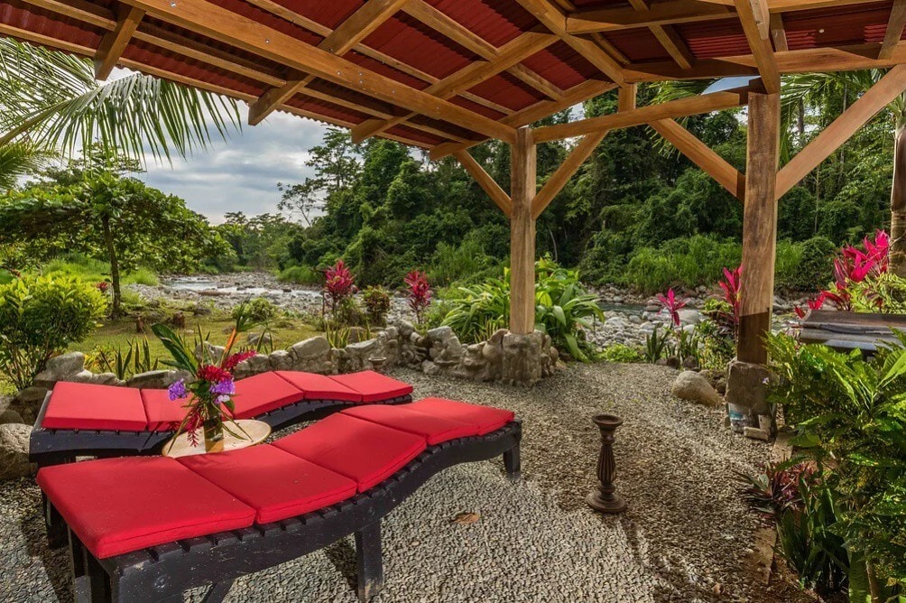 A covered patio area is set beside the river, featuring a series of red lounge chairs arranged around a circular table. Tropical plants surround the space, alongside decorative stones, creating a serene environment for relaxation with views of the flowing water and lush greenery.