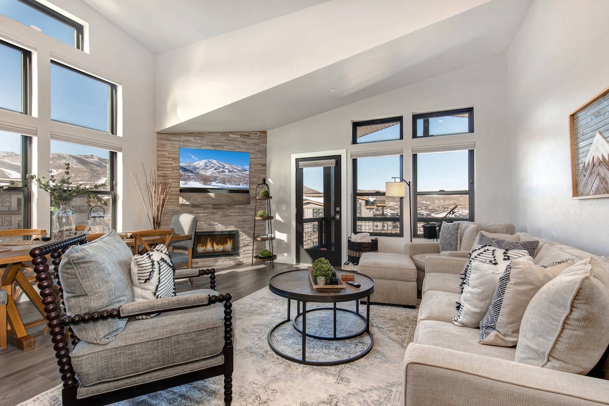 A bright living space features large windows that provide mountain views and natural light. Comfortable seating includes a neutral sofa and armchair, arranged around a coffee table. A stone fireplace is positioned beneath a mounted TV, adding a cozy element to the contemporary decor.