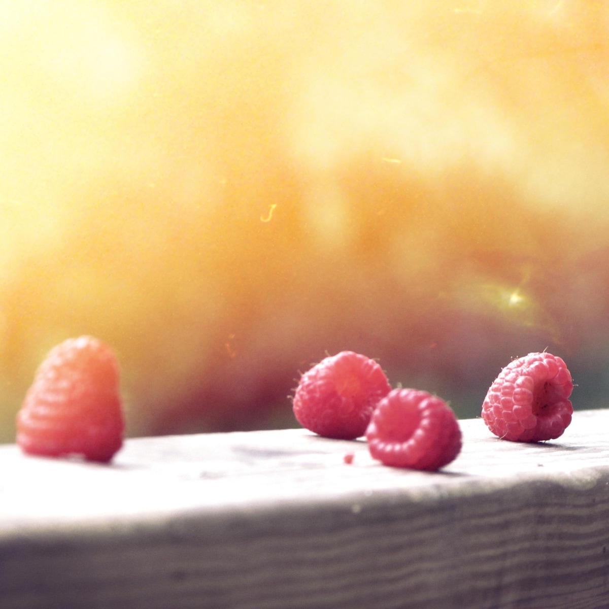 Raspberries are scattered on a weathered wooden surface. The soft focus background features warm, blurred colors, creating a serene atmosphere. Natural light enhances the vibrant red hues of the fruit.