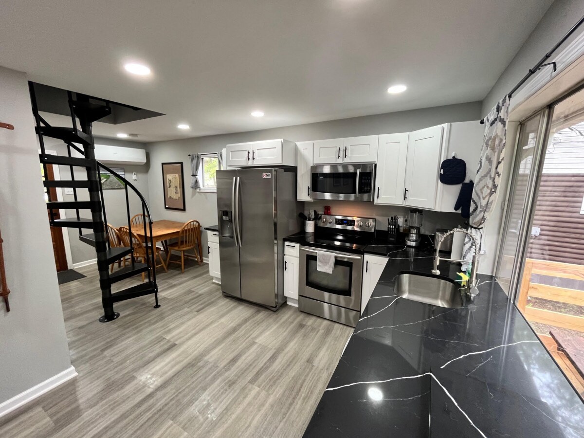 The kitchen is equipped with modern stainless steel appliances, including a refrigerator and microwave. A dark countertop contrasts with white cabinets. A spiral staircase leads to the loft area, while a dining table for four is positioned nearby. Natural light enters through a sliding door.