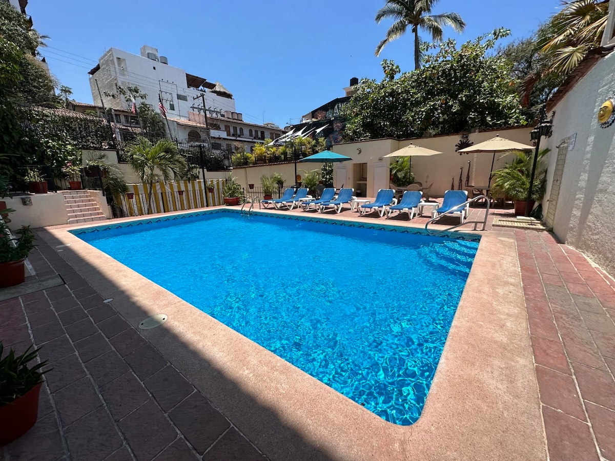 A large shared pool is situated in a sunlit courtyard, surrounded by lush greenery and shaded by umbrellas. Several lounge chairs are arranged along the pool’s edge, offering a relaxing space for guests. The vibrant blue water invites leisurely swims.