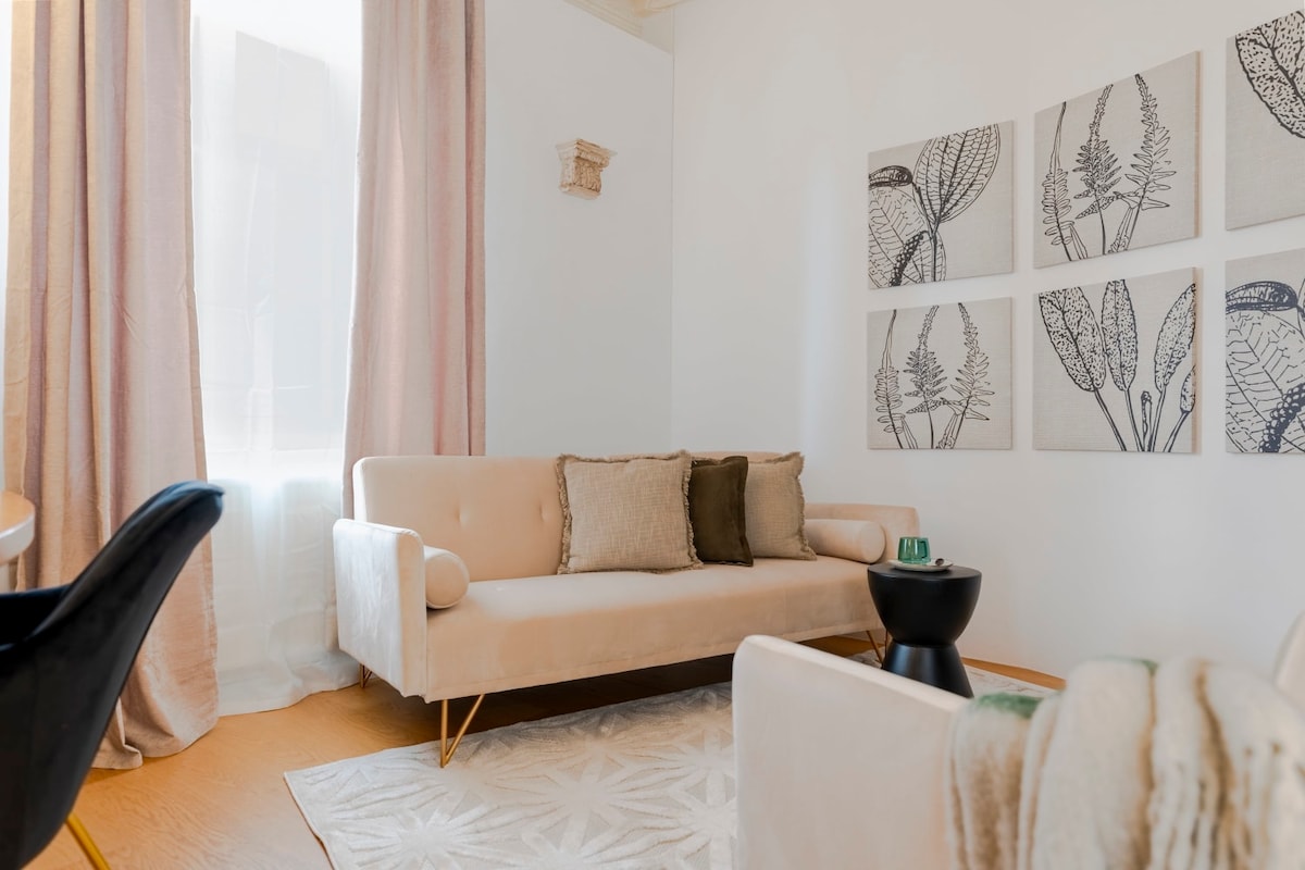 A light-filled living area showcases a soft, neutral sofa adorned with decorative cushions. A small black side table is positioned next to the sofa. Four patterned wall hangings are displayed above the sofa, adding visual interest to the space.