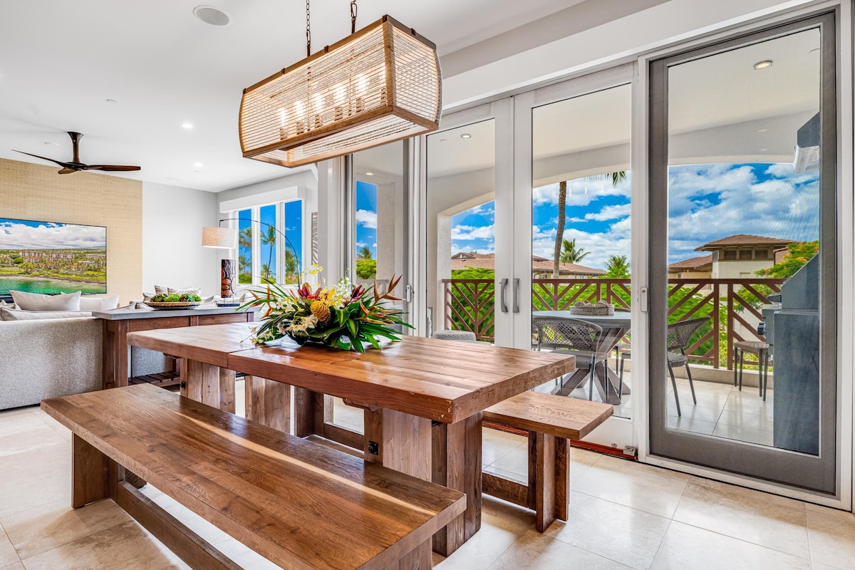 A spacious wooden dining table with bench seating is illuminated by a rectangular pendant light. Glass sliding doors frame a picturesque garden view, allowing natural light to fill the area. A tasteful floral arrangement adds a touch of color to the table, enhancing the inviting setting.