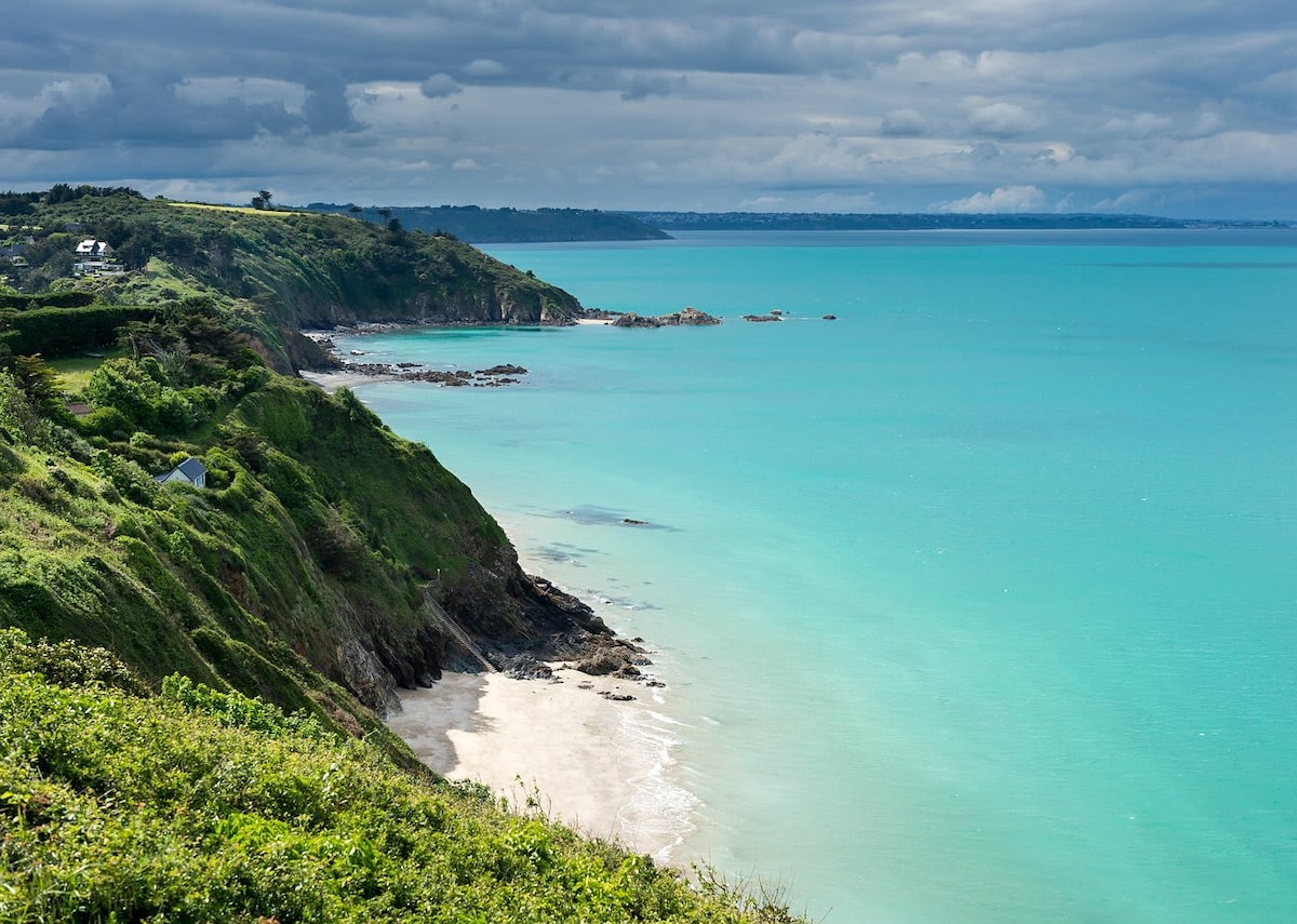 A coastal view captures the serene shoreline where turquoise waters meet sandy beaches. Cliffs are adorned with greenery, and gentle waves caress the rocky coastline under a partly cloudy sky. The landscape invites a sense of tranquility and connection with nature.