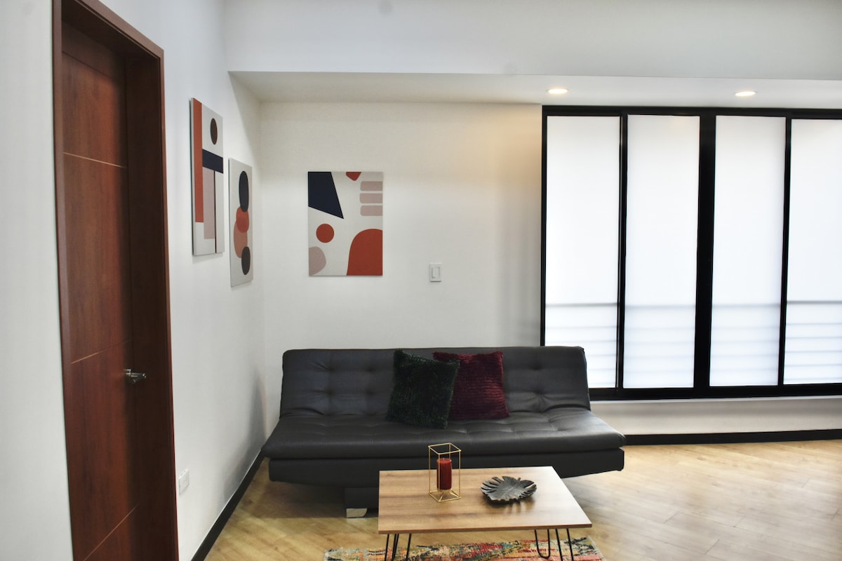 A modern living area is presented, featuring a dark sofa with plush cushions and a low wooden coffee table. Two abstract wall art pieces add color to the bright walls, while large windows fill the space with natural light, framed by light-colored blinds.
