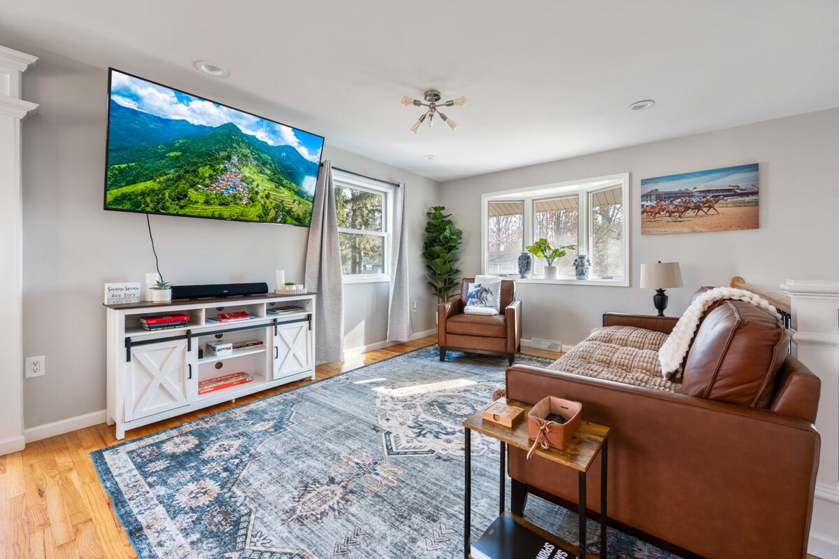 Gran televisor de pared, acogedor sofá de cuero y consola multimedia rústica con puerta de granero. La alfombra con estampado azul ancla la habitación. Grandes ventanales, vegetación y arte con temática de caballos añaden encanto. ¡Ideal para descansar o noches de cine!