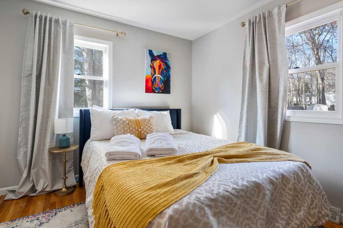 Dos grandes ventanales cubiertos con cortinas de color gris suave inundan la habitación de luz. La vibrante pintura de caballos ancla la pared sobre la cabecera azul marino. Ropa de cama fresca, manta acogedora y mesa auxiliar moderna completan esta escapada tranquila.