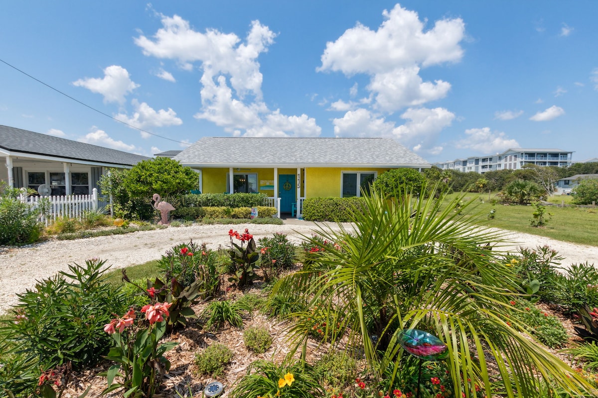 A vibrant yellow cottage is framed by lush greenery and colorful flowers. The inviting front porch is highlighted by a blue door. A circular driveway leads up to the entrance, creating an accessible and welcoming approach.