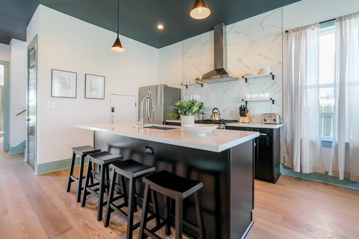 The kitchen features modern design elements, including a spacious island with seating for four. Sleek black cabinetry complements stainless steel appliances, and a marble backsplash adds a touch of elegance. Natural light filters through large windows, enhancing the bright and airy atmosphere.