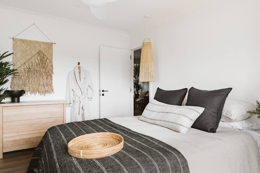 A cozy bedroom is presented, featuring a spacious bed adorned with dark and light-colored pillows. A woven throw and a decorative tray are placed atop the bed. Natural elements are showcased with a rattan pendant light and a wooden dresser, enhancing the calming atmosphere.