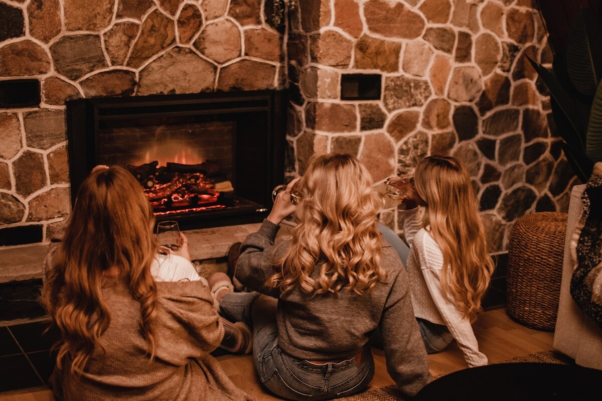 Three individuals are seated on the floor with their backs to the camera, enjoying drinks in front of a stone fireplace. The cozy ambiance is enhanced by the warm glow of the fire, with rustic decor visible in the background.