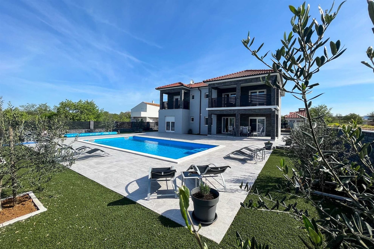 The villa's exterior showcases a large heated pool surrounded by modern sun loungers. Lush greenery and Mediterranean trees enhance the outdoor space, while the spacious structure features multiple balconies and an inviting layout that encourages relaxation and outdoor enjoyment.