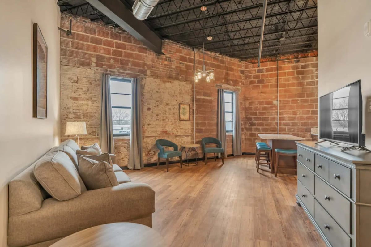 An open living area showcases exposed brick walls, contributing to the loft's industrial charm. Soft seating complements a light wooden floor, while two large windows provide natural light. A television is positioned on a light-colored dresser, with a dining table in the background.