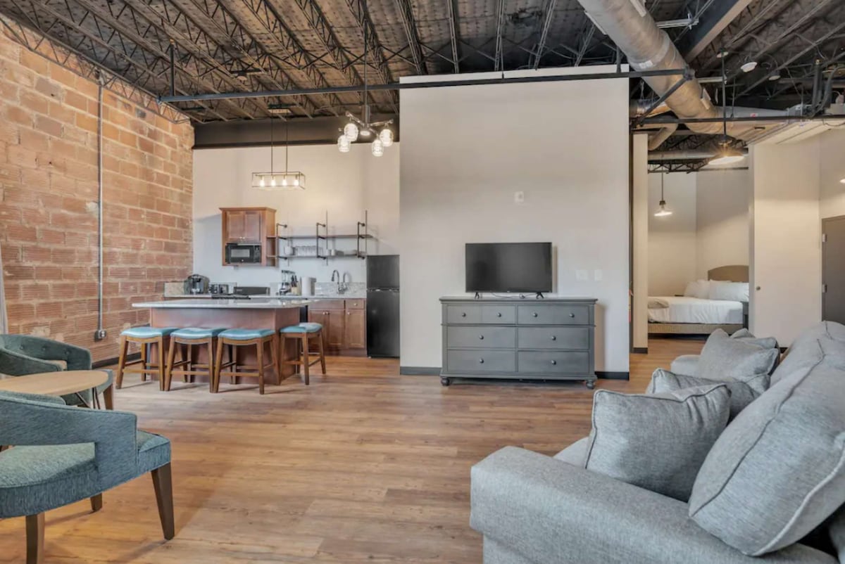 An open-concept living area showcases a blend of exposed brick and modern furnishings. The kitchen area features sleek cabinets and a breakfast bar with four stools. A comfortable seating arrangement is visible in soft grey upholstery. Light hardwood flooring adds warmth throughout the space.