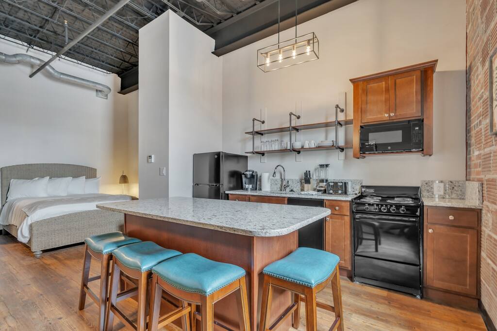 An open-concept loft reveals a modern kitchen with granite countertops and stainless steel appliances. A spacious kitchen island with three blue stools invites casual dining. The background features a cozy sleeping area with a large bed, enhancing the loft's welcoming atmosphere.
