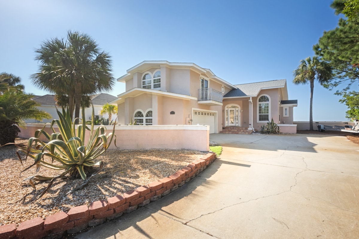The exterior of a spacious two-story home is showcased with a circular driveway lined with smooth concrete. Lush palm trees and decorative plants are visible in the landscaped front yard, complemented by the soft hues of the home's facade under a clear blue sky.