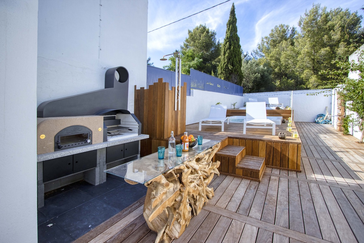 An outdoor terrace features a distinctive wooden table and seating area, with a stone barbecue and pizza oven nearby. Sun loungers are positioned around the space, which is surrounded by lush greenery, creating a serene environment for relaxation and dining.