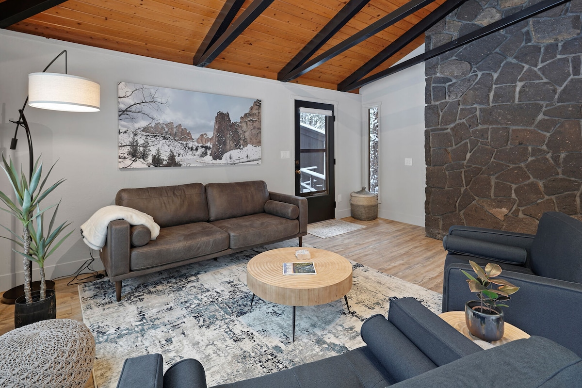 A modern living room showcases a dark couch positioned across from a round wooden coffee table. A cozy area rug covers the floor, and a large stone wall adds texture to the space. Natural light enters through a window, illuminating the room's rustic decor.