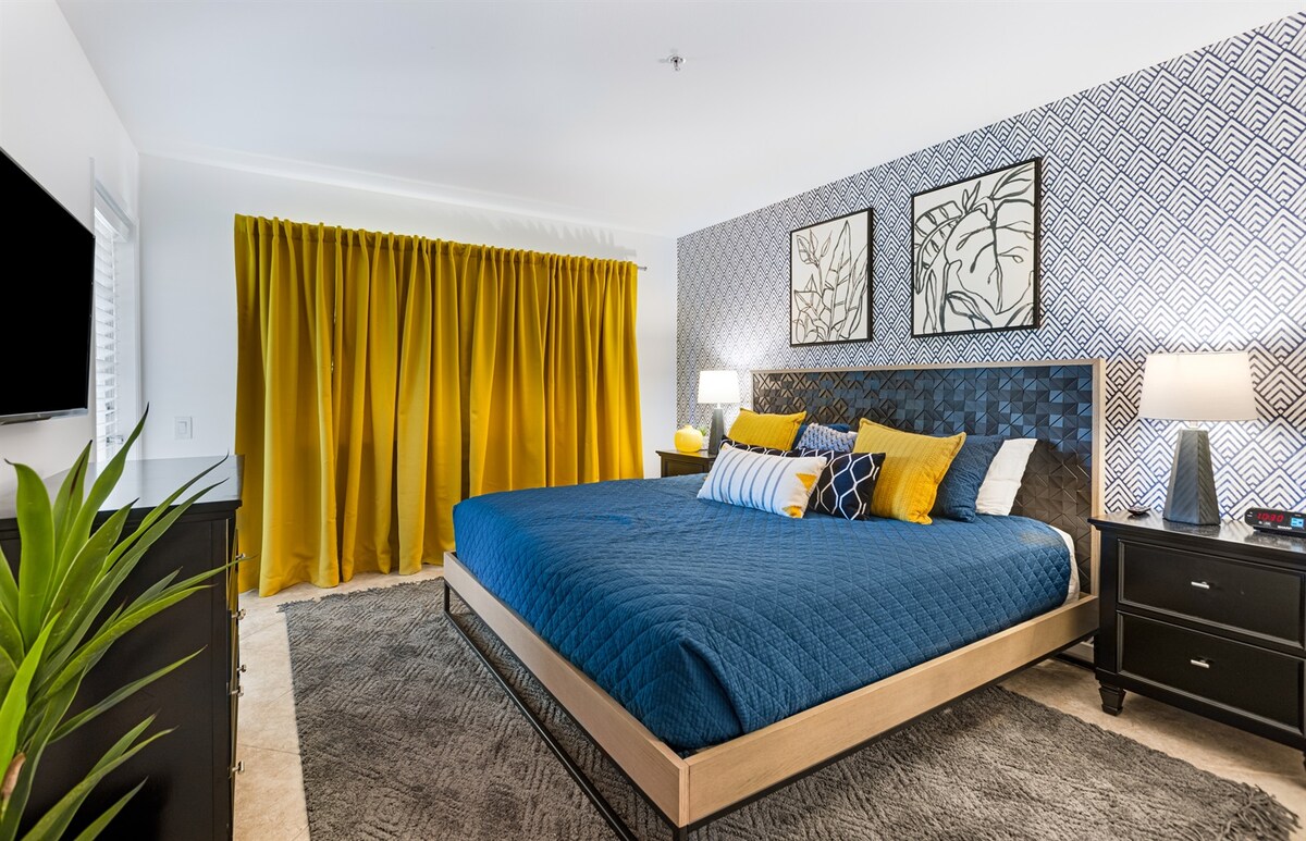 A spacious bedroom is highlighted by a large king bed adorned with a blue quilt and decorative pillows in various vibrant shades. Colorful artwork is displayed above the bed. Floor-to-ceiling yellow curtains frame a window, while a black dresser and nightstands provide functional elegance.