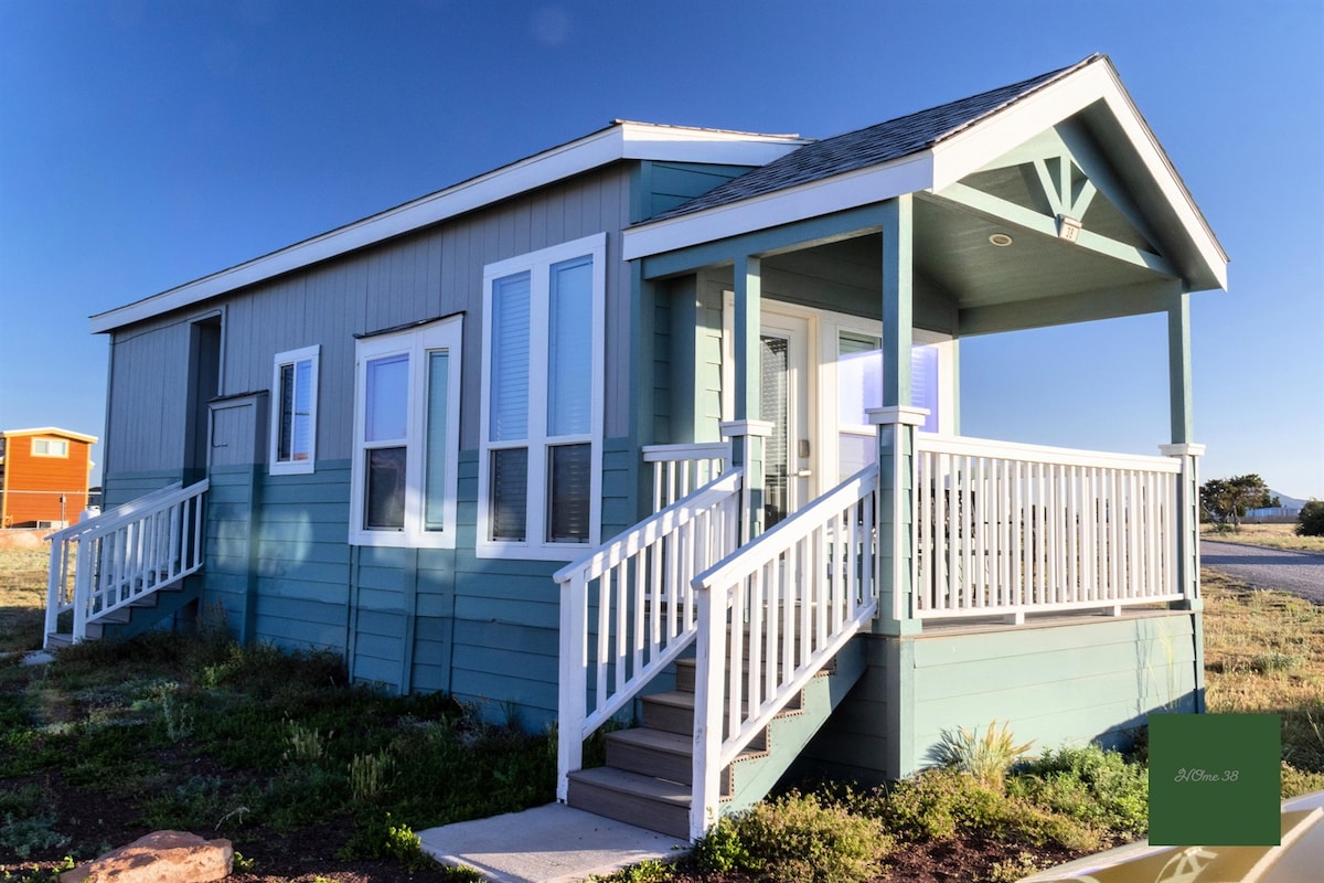 The exterior of a tiny home is shown, featuring a light blue wooden facade with white trim. A welcoming porch with white railings and steps leads up to the front entrance. Large windows allow ample natural light, highlighting the home's cheerful design in a quiet outdoor setting.