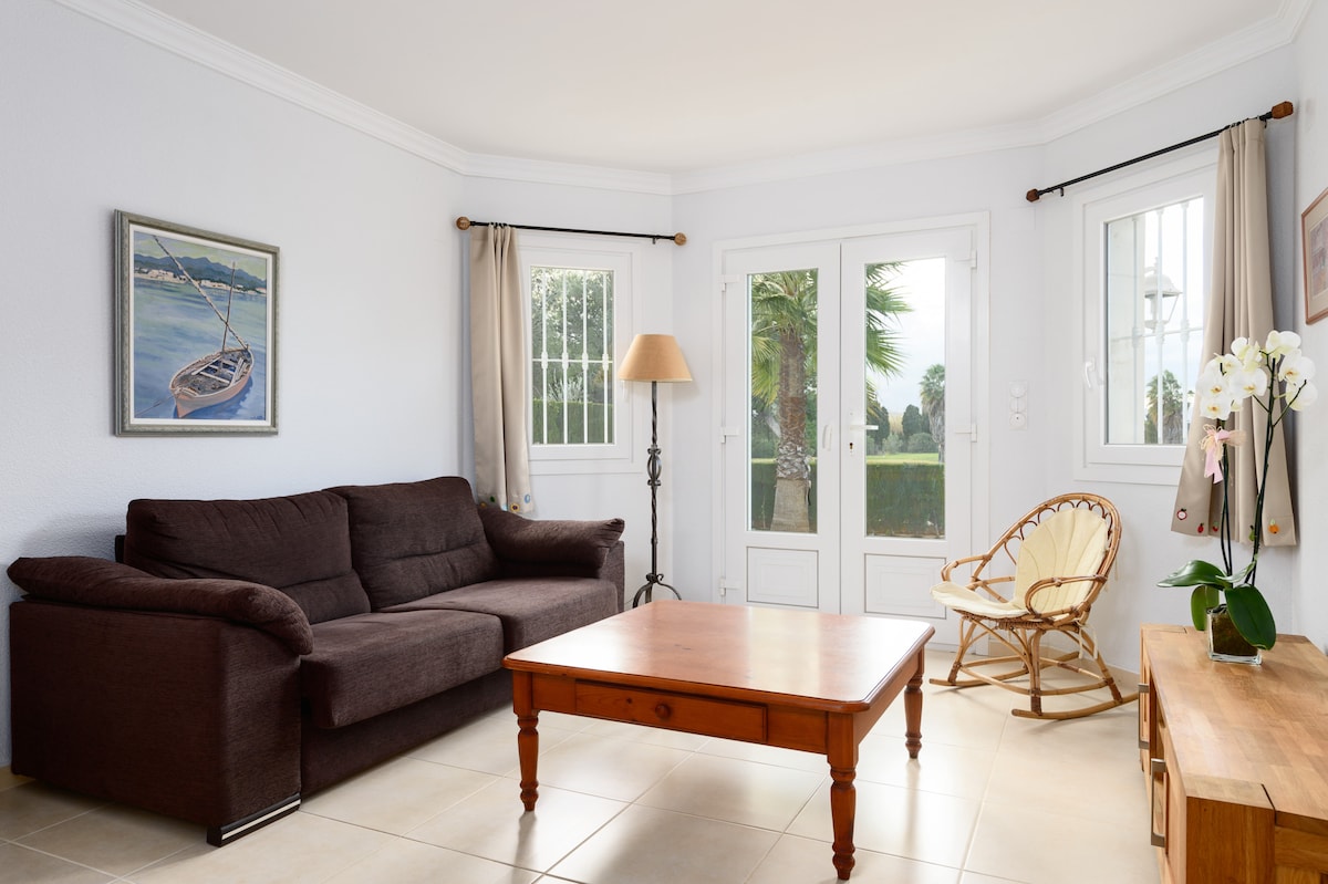 A cozy living area features a brown sofa and a wooden coffee table, illuminated by natural light from two large windows. A rattan armchair adds to the seating options, while a floor lamp provides additional warmth. A piece of artwork depicts a boat on water.