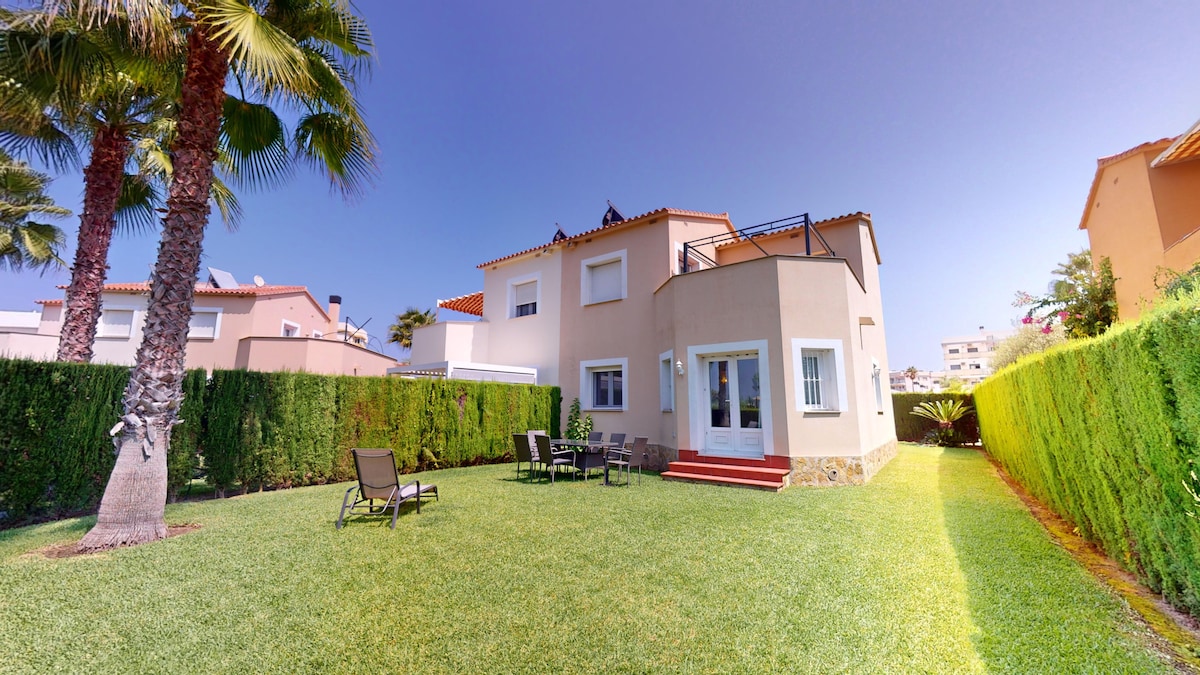 The semi-detached villa is set within a private garden, surrounded by green hedges. Lounge chairs and a dining table are positioned on the grassy area, providing an inviting outdoor space. The exterior features a light-colored facade with multiple windows, complemented by palm trees.