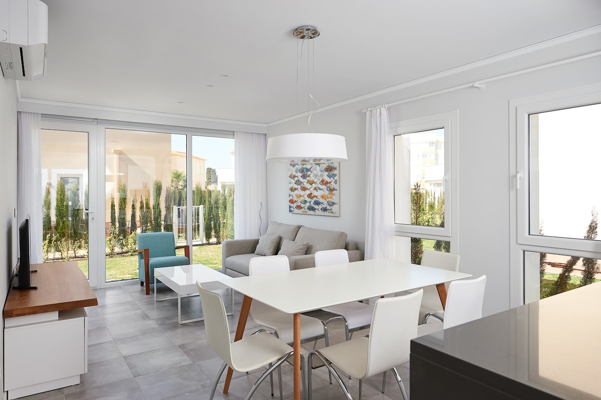 An open-plan living area features a comfortable seating arrangement with a light grey sofa and colorful artwork on the wall. A white dining table is surrounded by four chairs, while large glass doors offer views of the private garden and allow natural light to fill the space.