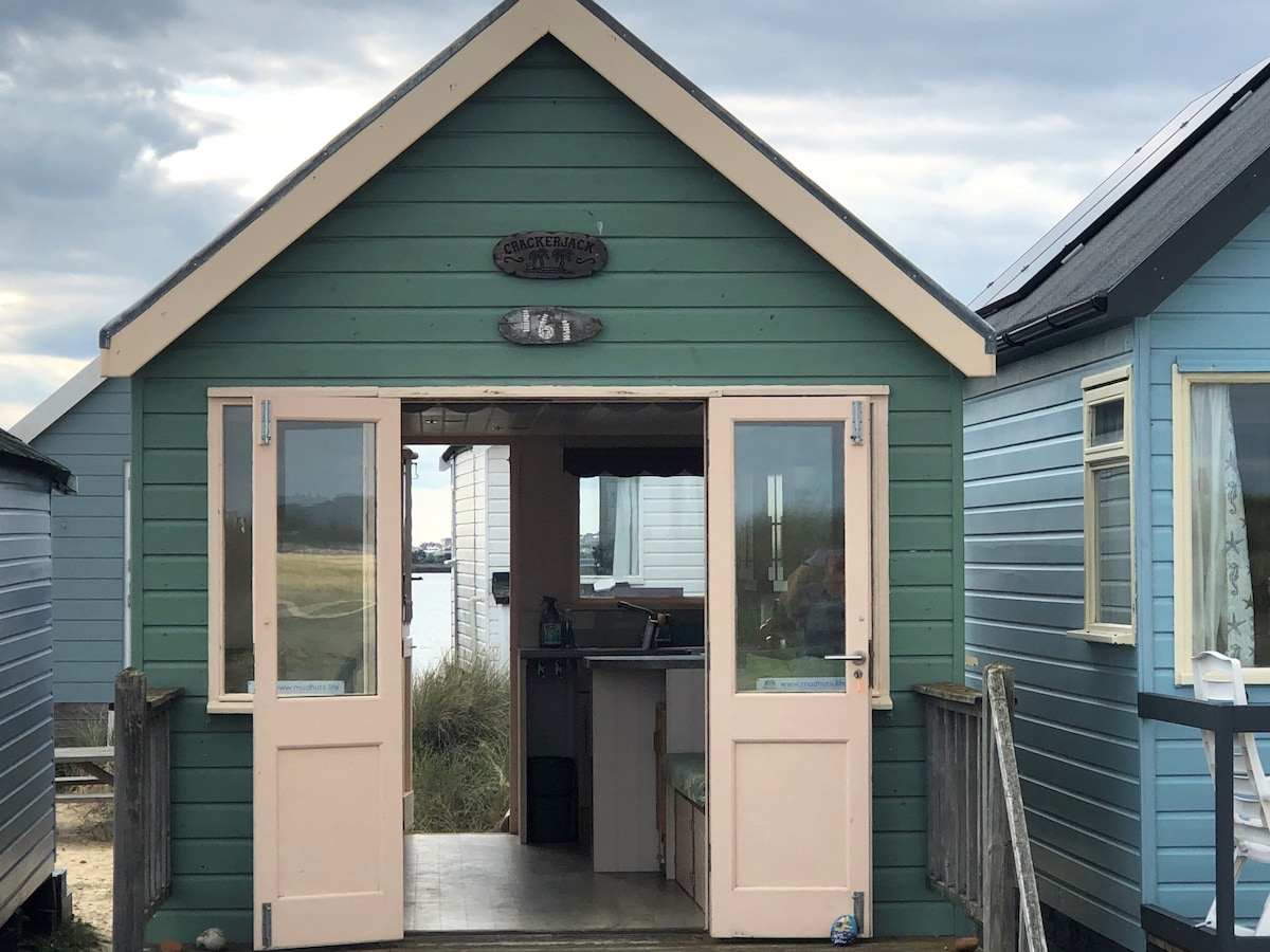 The exterior of Crackerjack hut is shown, featuring a green facade with light pink doors. A sign with the number '161' is prominently displayed above the entrance. The surrounding area includes neighboring beach huts, while glimpses of the landscape and water can be seen through the open doors.