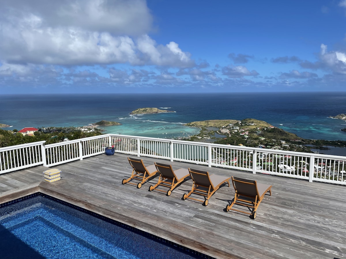An outdoor deck features a sparkling swimming pool bordered by four chaise lounges, overlooking an expansive view of the Caribbean Sea and the surrounding islands. The sky is partially cloudy, highlighting the coastal landscape in the distance.