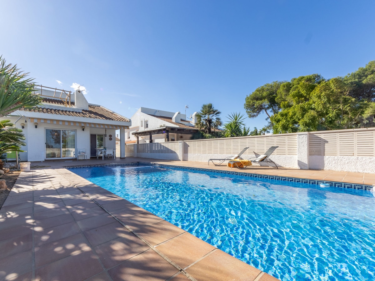 A spacious swimming pool is set within a well-maintained outdoor area, featuring clear blue water and sun loungers positioned nearby. A single-story house, partially visible, is framed by palm trees and greenery against a bright blue sky.