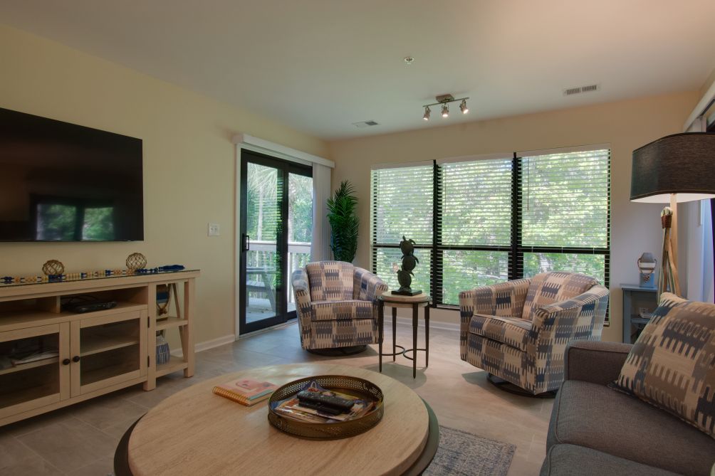 The living room features contemporary furnishings, including two patterned armchairs and a cozy sofa. Oversize windows allow natural light to fill the space, while a coffee table and a TV unit add functionality. Lush greenery can be seen through the windows, enhancing the view.