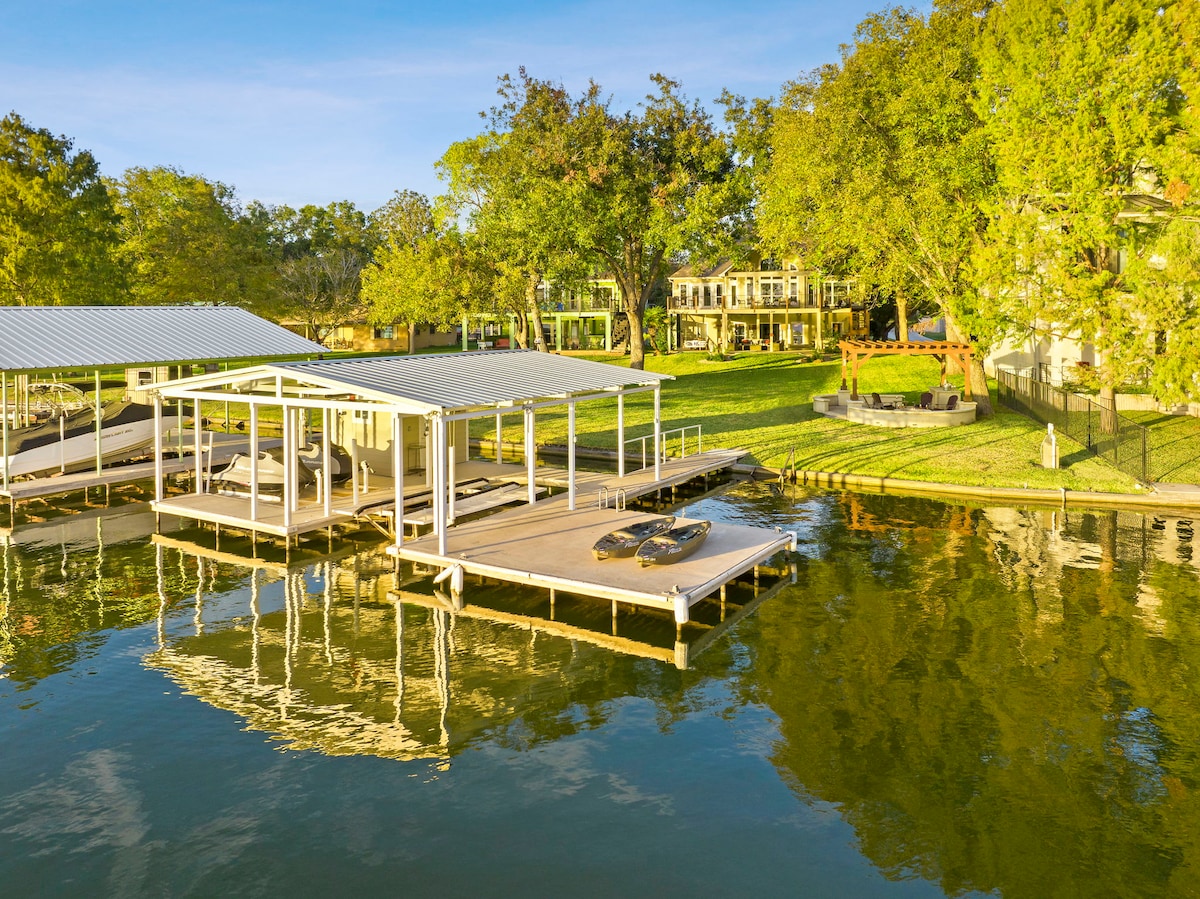 The waterfront area features a spacious dock with boat lifts and a covered boat slip. Surrounding greenery provides a natural backdrop. Reflections of the trees and sky can be seen on the water's surface, enhancing the tranquil lakeside setting.
