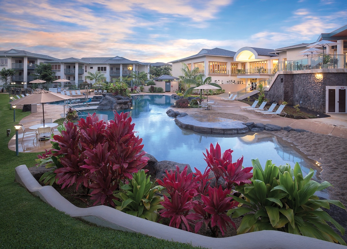 A serene resort setting features a large pool surrounded by lush tropical plants and comfortable lounge chairs. Umbrellas provide shade near the water's edge, while the soft colors of the sky reflect on the pool's surface, creating a tranquil atmosphere for relaxation.