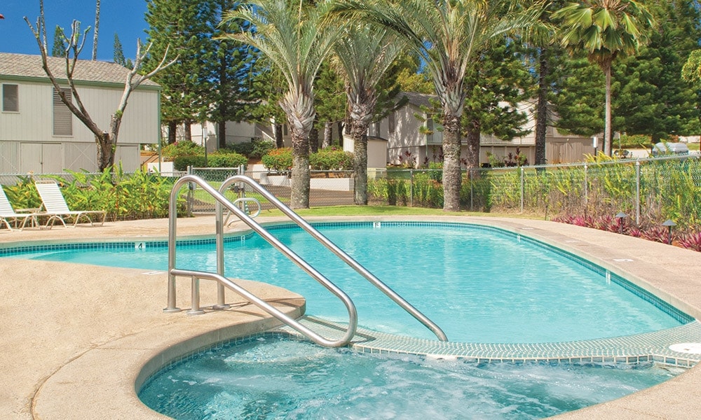 An outdoor swimming pool features crystal clear water, bordered by a smooth concrete deck. A hot tub is adjacent to the pool, equipped with a stainless steel railing for easy access. Palm trees and lush greenery surround the area, providing a natural backdrop.