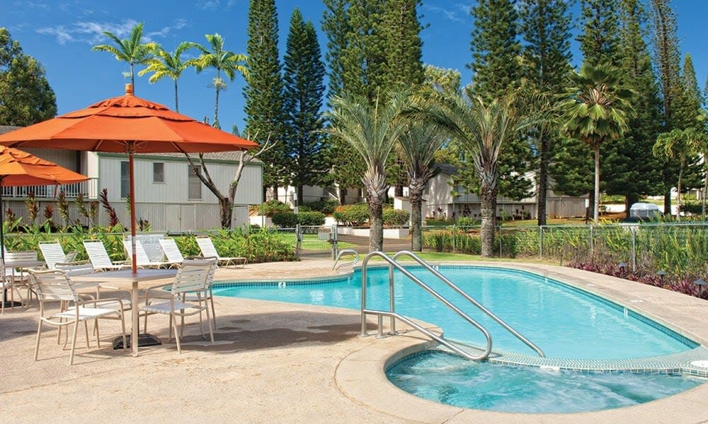 An outdoor swimming pool is surrounded by lush greenery and tropical palm trees. Several lounge chairs with orange umbrellas are positioned nearby, offering shaded seating. A small building is visible in the background, contributing to the resort setting.
