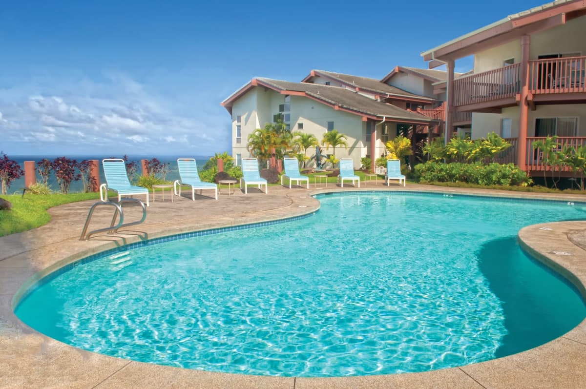 An outdoor swimming pool is surrounded by lounge chairs and vibrant landscaping. Palm trees and tropical plants are visible, enhancing the serene environment. The building's balconies overlook the pool, and clear blue skies frame the scene.
