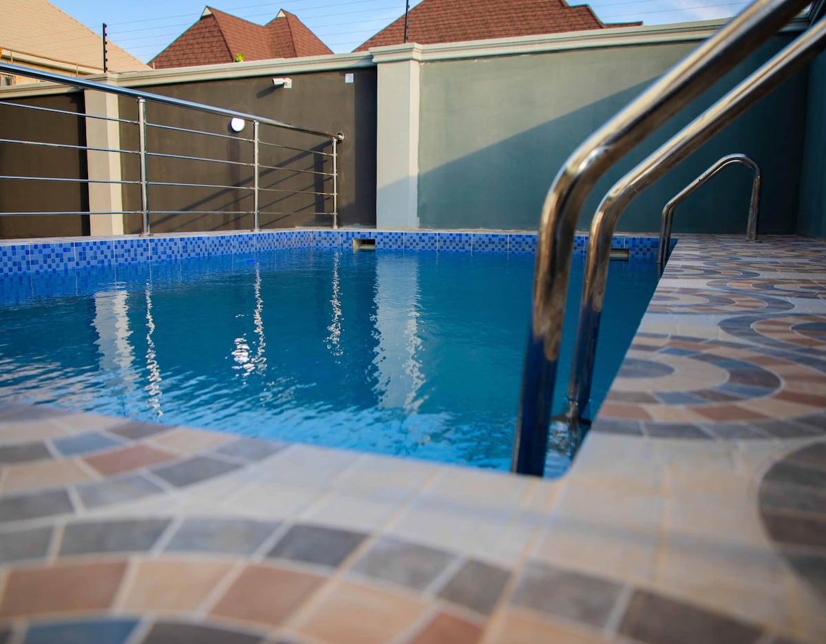 A clear and inviting swimming pool is surrounded by modern tiled flooring, featuring a gentle entry ramp and stainless steel handrails. The water's surface reflects the surrounding architecture and sky, creating a serene setting within the compound.
