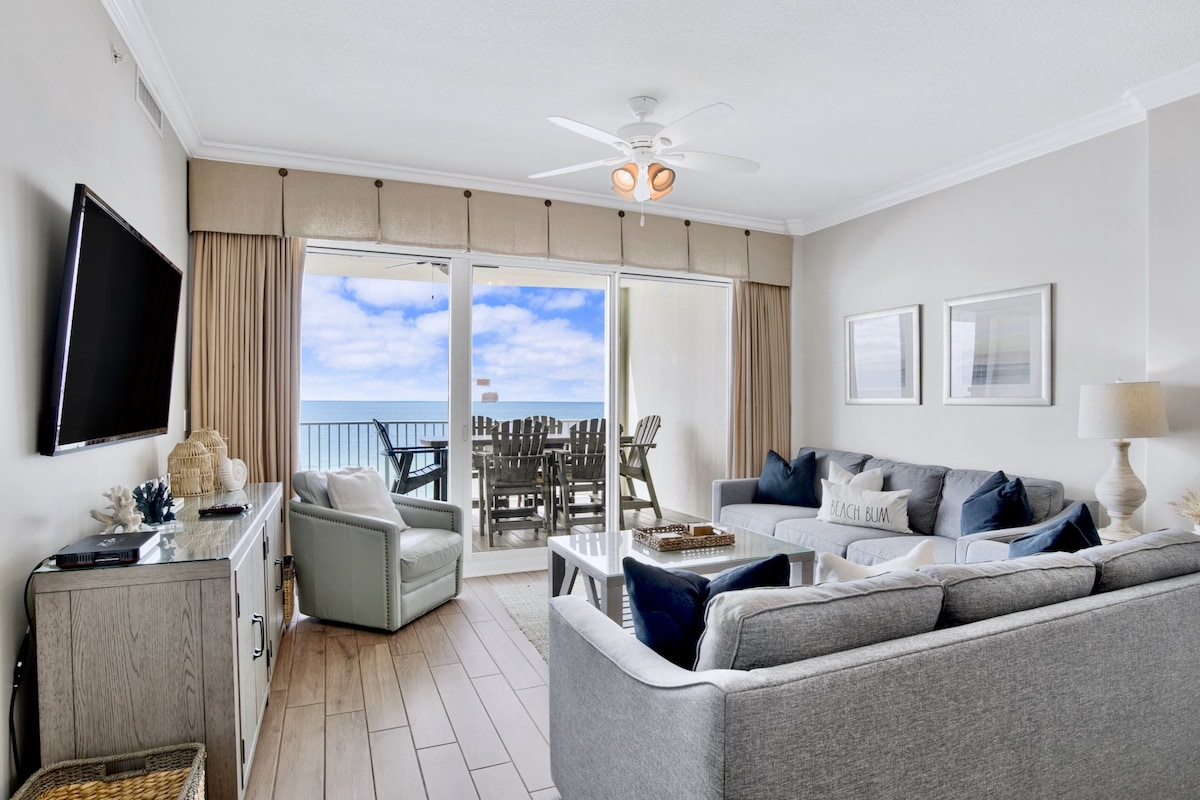 A spacious living area features soft grey seating arrangements and a ceiling fan. Natural light fills the room through large sliding glass doors that open to a private balcony with outdoor seating. A flat-screen TV is mounted on the wall, complementing the neutral decor.