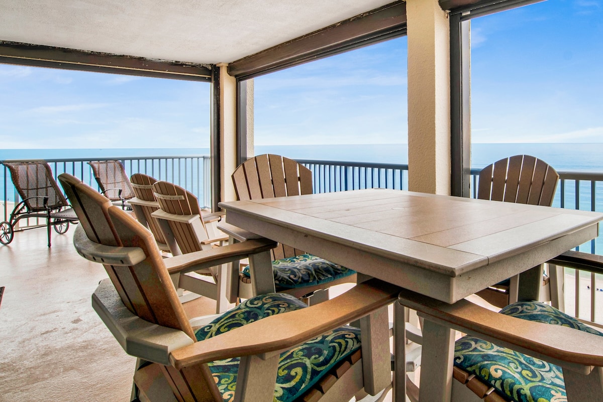 A spacious balcony features a dining table surrounded by wooden chairs with decorative cushions. The ocean can be seen in the background, framed by large glass sliding doors. Comfortable lounge chairs are positioned to the side, providing a relaxing space to enjoy the views.