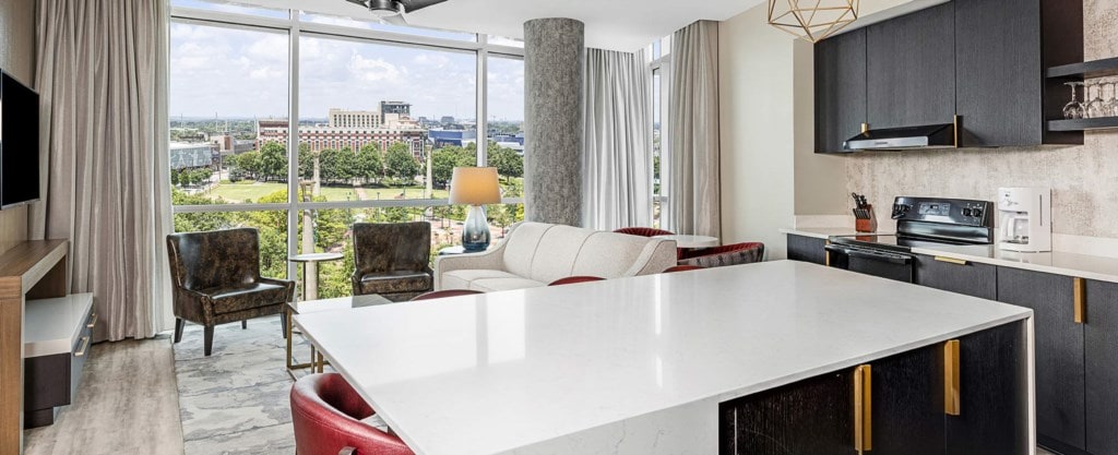 A modern living area is highlighted by large windows offering views of the cityscape. A spacious white countertop divides the kitchen space from the living area, where a cream-colored sofa and two dark armchairs are positioned. Natural light fills the room through the sheer curtains.
