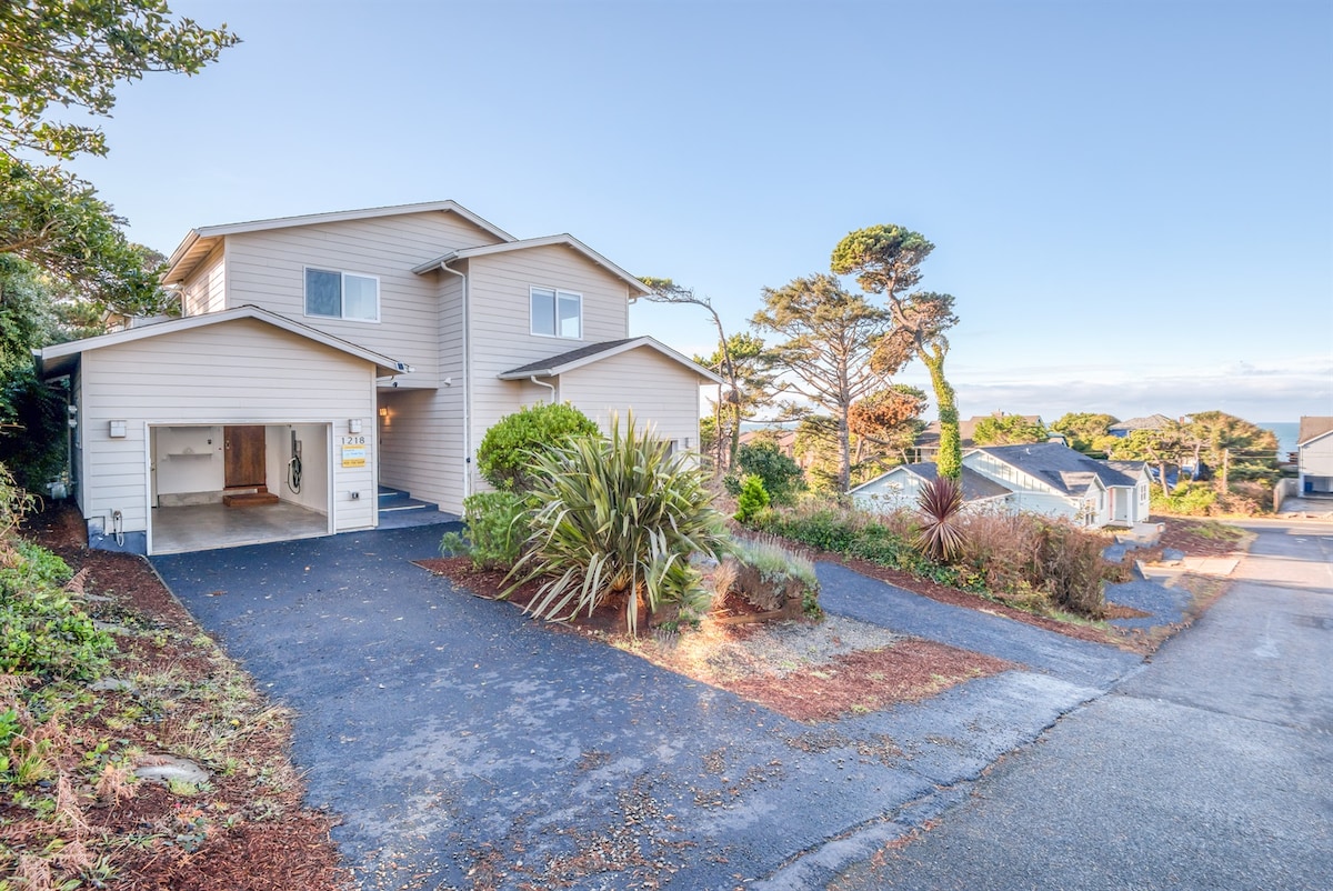 A two-story duplex is shown, featuring a light-colored exterior and a welcoming front entry. The driveway is paved, leading to a garage on the left. Surrounding greenery includes shrubs and trees, contrasting with the clear blue sky in the background.