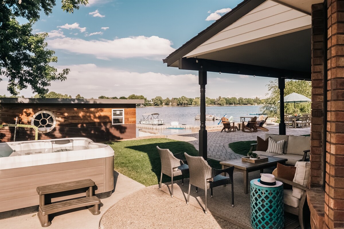 A serene outdoor space features a private hot tub alongside patio seating arranged under a shaded roof. A spacious green lawn leads towards a tranquil lake, with lounge chairs and a private dock visible in the background, inviting relaxation and enjoyment of the picturesque surroundings.