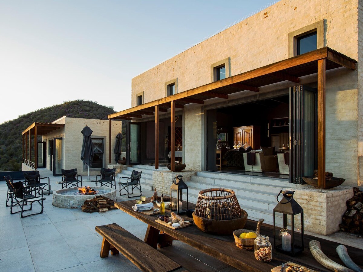 An outdoor dining area features a long wooden table, surrounded by chairs and lanterns. The space is complemented by a built-in fire pit and a backdrop of the lodge's modern architecture, highlighted by expansive windows and shaded patio spaces. Scenic mountains rise in the distance.
