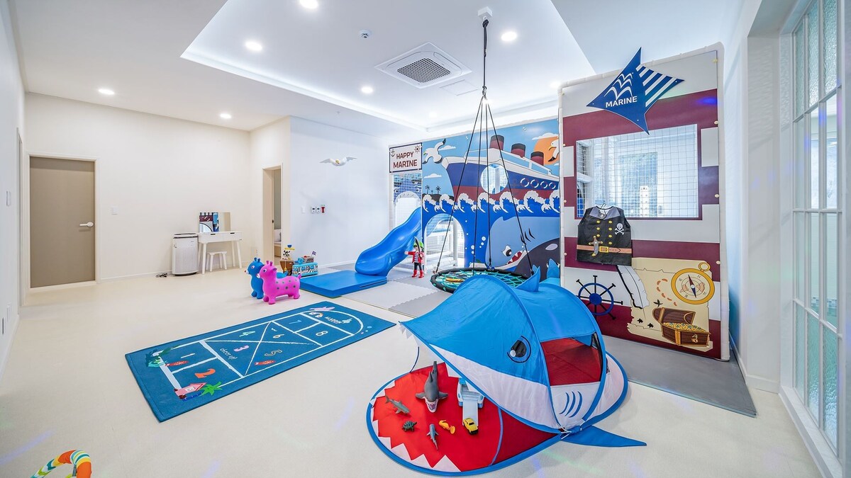 A spacious children's playroom is designed with bright colors and playful decor. A blue slide is featured alongside a tent shaped like a shark. Various toys are scattered on a soft carpet, and a themed mural depicting a ship adds an adventurous touch to the inviting space.