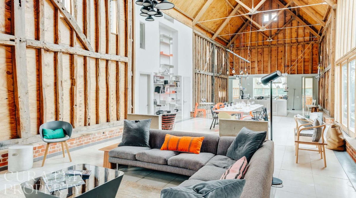 The spacious open-plan living area features high wooden beams and large windows allowing natural light to flow in. A comfortable sectional sofa with colorful cushions is positioned in front of a sleek coffee table, while a dining area is visible in the background, elegantly furnished for group gatherings.